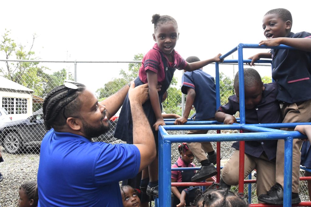 Hakeem Atkinson, Member Relations Supervisor at JN Group, assists a student at Chapel Hill Basic School in climbing the monkey bar, one of the pieces of equipment donated to the school.